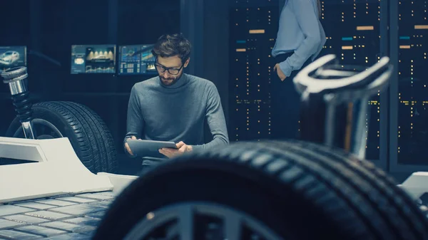 汽车设计工程师坐在混合动力汽车底盘平台原型旁边,使用平板电脑进行设计增强。带悬架、车轮、发动机和蓄电池的车身框架设施 — 图库照片