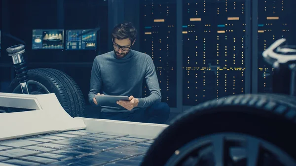 汽车设计工程师坐在混合动力汽车底盘平台原型旁边,使用平板电脑进行设计增强。带悬架、车轮、发动机和蓄电池的车身框架设施 — 图库照片