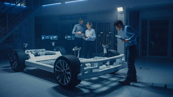 Diversos engenheiros de automóveis conversando enquanto trabalhava no protótipo de chassi de plataforma de carro elétrico. No conceito de instalação de inovação automotiva, o quadro do veículo inclui pneus, suspensão, motor e bateria — Fotografia de Stock