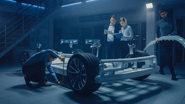電気自動車プラットフォームのシャーシに取り組む自動車エンジニアのチームは、対策を講じ、3D Cadソフトウェアと協力し、効率を調査します。車輪、エンジンおよび電池が付いている車のフレーム. — ストック写真