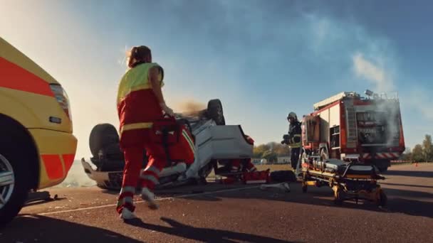 Sanitariusze i strażacy przybywają na miejsce wypadku samochodowego. Profesjonaliści ratują poszkodowaną ofiarę uwięzioną w Rollover Vehicle poprzez ich wydobycie, udzielanie pierwszej pomocy i gaszenie pożaru — Wideo stockowe