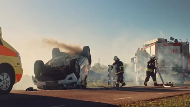 Sanitariusze i strażacy przybywają na miejsce wypadku samochodowego. Profesjonaliści ratują poszkodowaną ofiarę uwięzioną w Rollover Vehicle poprzez ich wydobycie, udzielanie pierwszej pomocy i gaszenie pożaru — Wideo stockowe