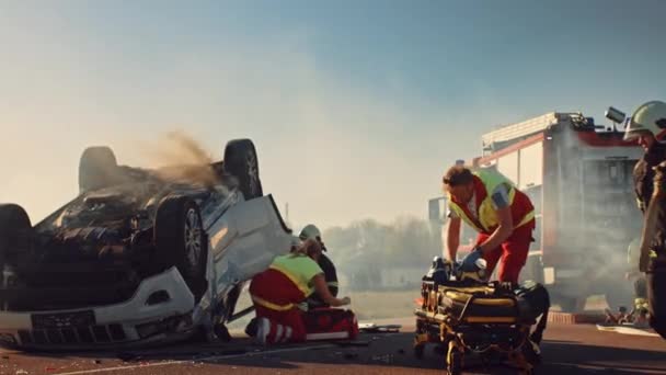 Am Unfallort: Sanitäter und Feuerwehrleute retten Verletzte, die im Fahrzeug eingeklemmt sind. Sanitäter benutzen Tragen, leisten Erste Hilfe. Feuerwehrleute greifen nach Ausrüstung. Zeitlupe — Stockvideo