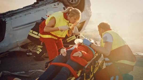 En la escena del accidente de tráfico: Equipo de rescate de bomberos saca a la víctima femenina del vehículo de vuelco, usan camillas con cuidado, la entregan a paramédicos que realizan primeros auxilios — Vídeo de stock