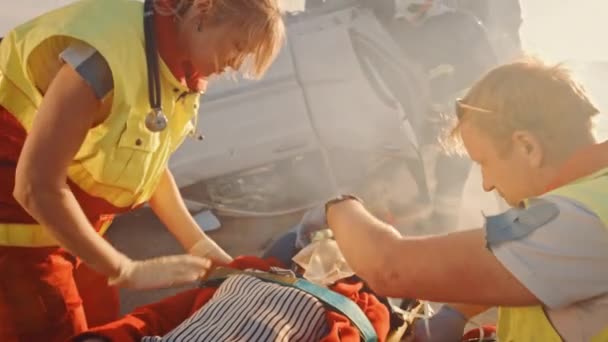 Am Unfallort: Sanitäter retten einem weiblichen Opfer, das auf Tragen liegt, das Leben. Sie hören einen Herzschlag, setzen eine Sauerstoffmaske auf und leisten Erste Hilfe. Feuerwehr löscht Brand — Stockvideo