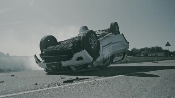 Příšerná dopravní nehoda Rollover Kouření a hořící vozidlo ležící na střeše uprostřed silnice po srážce. Denní havárie s poškozeným autem. Shot In Black and White — Stock video