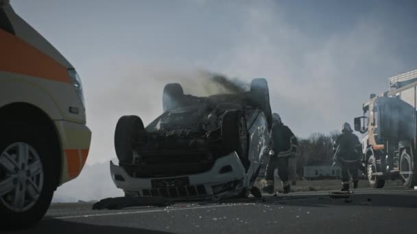 Sanitäter und Feuerwehrleute treffen am Unfallort ein. Profis retten verletztes Opfer, das in Rollover-Fahrzeug eingeklemmt ist, indem sie es befreien, Erste Hilfe leisten und Feuer löschen — Stockvideo