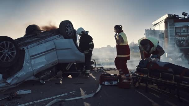 Op de auto crash verkeersongeval scene: paramedics geven eerste hulp zuurstofmasker aan vrouwelijke slachtoffer van het ongeval. Brandweerlieden blussen vuur en gebruiken hydraulische frees om andere passagiers te vrijen — Stockvideo