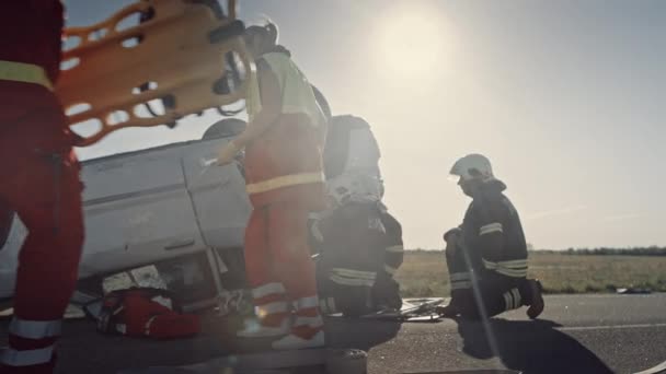 自動車事故交通事故:救急隊員と消防士は、ロールオーバー車両に閉じ込められた乗客を救出する計画を立てます。メディックは応急処置装置と緊急ストレッチャーを準備します。消防士は油圧カッター拡散機を使用する — ストック動画