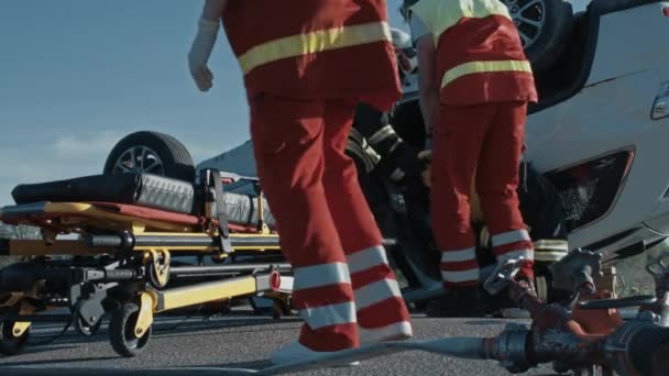 Am Unfallort: Rettungsteam der Feuerwehr zieht weibliches Opfer aus Überschlagfahrzeug, setzt Tragen vorsichtig ein, übergibt es an Sanitäter, die Erste Hilfe leisten. niedriger Winkel — Stockvideo