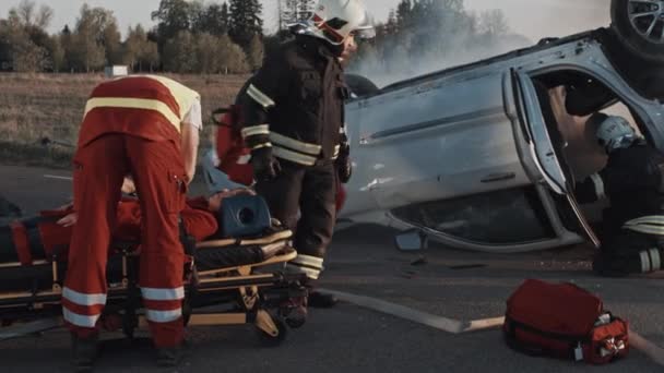 自動車事故交通事故:救急隊員と消防士が負傷した犠牲者を救出。メディックはショックを受けた女性の乗客ストレッチャーに応急処置を与えます。消防士は、車両を開くために油圧カッター拡散機を使用します — ストック動画