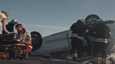 Araba kazasında trafik kaza sahnesinde: sağlık görevlileri sedye üzerinde yatan bir yolcu yaşam kaydedin. Onlar bir Heartbeat dinleyin, oksijen maskesi kullanın, kurulum Drop Counter. İtfaiyeciler araba açmak için kesiciler kullanın