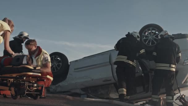 Am Unfallort: Sanitäter retten einem auf Tragen liegenden Beifahrer das Leben. Sie hören einen Herzschlag, benutzen eine Sauerstoffmaske, richten einen Fallzähler ein. Feuerwehr öffnet Auto mit Fräsern — Stockvideo