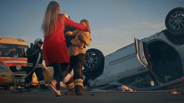 Dappere brandweerman draagt gewond jong meisje naar veiligheid waar ze herenigd met haar liefhebbende moeder. Op de achtergrond Auto crash Verkeersongeval Courageous Paramedici en brandweerlieden redden levens — Stockvideo