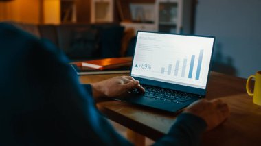 Omuz shot Over: genç adam Desk içinde oturan Istatistik ve Infographics gösterir bir laptop çalışır. Gece geç saatlerde onun oturma odası adam bilgisayar kullanır.
