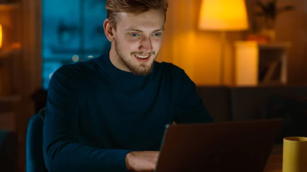 Bonito sorridente homem trabalha em um laptop enquanto sentado em sua mesa em casa. Retrato de um jovem freelancer funciona no computador em sua acolhedora sala de estar com iluminação noturna quente no fundo . — Fotografia de Stock