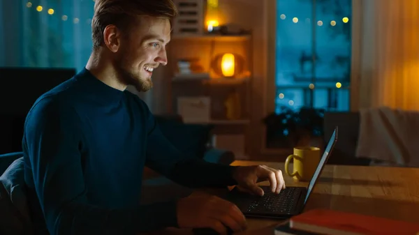 Βράδυ στο σπίτι: πορτρέτο του όμορφος άνθρωπος κάθεται στο γραφείο του δουλεύοντας σε ένα φορητό υπολογιστή. Ο χαμογελαστός ανεξάρτητος δουλεύει στον υπολογιστή. Στιγμιότυπο πλευρικής προβολής. — Φωτογραφία Αρχείου