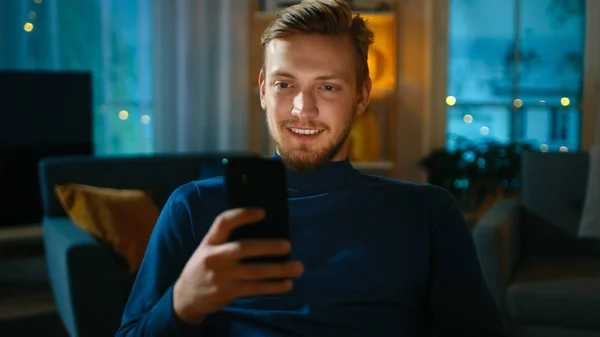 Handsome Man Relaxes at Home Uses Smartphone. In the Background Cozy Living Room in the Evening with Warm Lights On.