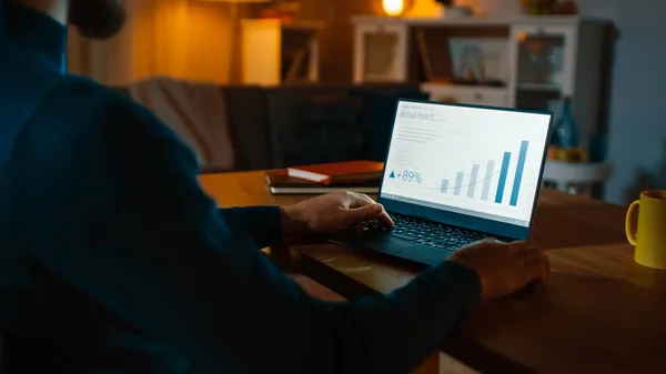 Abend zu Hause: Der Mann arbeitet an einem Laptop, der eine statistische Infografik zeigt. gemütliches Wohnzimmer mit warmem Licht. — Stockfoto