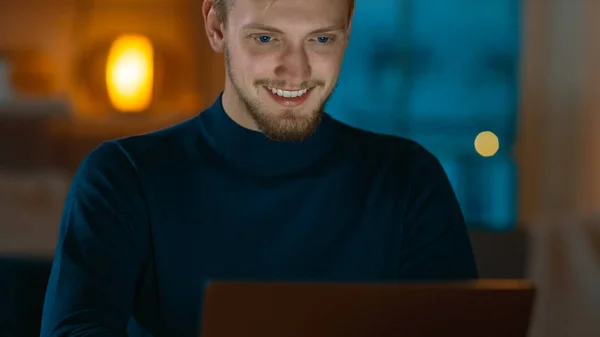 Noite em casa: retrato de um homem bonito sentado em sua mesa trabalhando em um laptop. Freelancer sorrindo Trabalhando no computador . — Fotografia de Stock