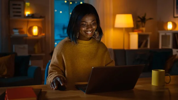 Πορτρέτο του όμορφη χαμογελαστή αφρικανική αμερικανική κορίτσι χρησιμοποιώντας laptop ενώ κάθεται στο γραφείο της στο σπίτι. Το βράδυ δημιουργική γυναίκα εργάζεται σε έναν υπολογιστή στο φιλόξενο σαλόνι της. — Φωτογραφία Αρχείου