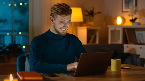 Улыбающийся красавчик работает на ноутбуке, сидя за своим столом дома. Портрет молодого фрилансера работает за компьютером в уютной гостиной с теплым вечерним освещением на заднем плане . — стоковое фото