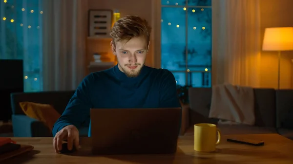 Βράδυ στο σπίτι: πορτρέτο του όμορφος άνθρωπος κάθεται στο γραφείο του δουλεύοντας σε ένα φορητό υπολογιστή. Χαμογελαστός ελεύθερος επαγγελματίας εργασίας στον υπολογιστή. — Φωτογραφία Αρχείου