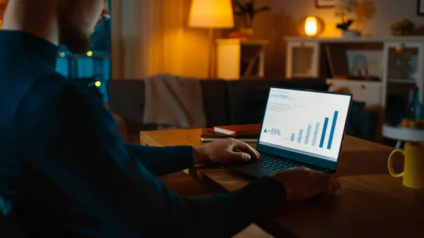 Evening at Home: Man Works on a Laptop Showing Statistical Infographic (en inglés). Acogedor salón con luces cálidas . —  Fotos de Stock