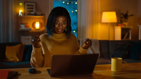 In de avond: portret van mooi glimlachend meisje met laptop terwijl ze thuis aan haar bureau zat, plotseling wint ze groot en viert ze haar overwinning met Yes gebaar. — Stockfoto