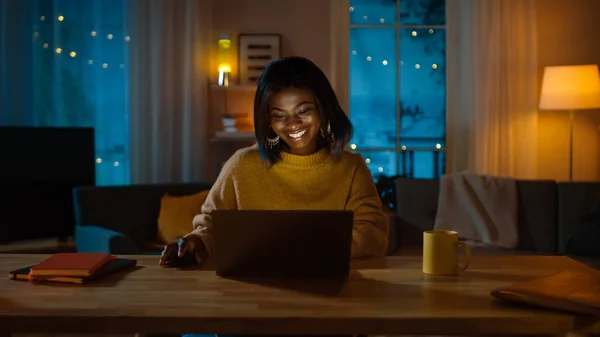 Retrato de una hermosa chica negra sonriente trabajando en una computadora portátil mientras está sentada en su escritorio en casa. Por la noche, la mujer creativa trabaja en una computadora en su acogedora sala de estar . —  Fotos de Stock