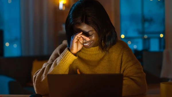 Πορτρέτο του όμορφη χαμογελαστή μαύρο κορίτσι δουλεύοντας σε ένα φορητό υπολογιστή, ενώ κάθεται στο γραφείο της στο σπίτι. Το βράδυ δημιουργική γυναίκα εργάζεται σε έναν υπολογιστή στο φιλόξενο σαλόνι της. — Φωτογραφία Αρχείου