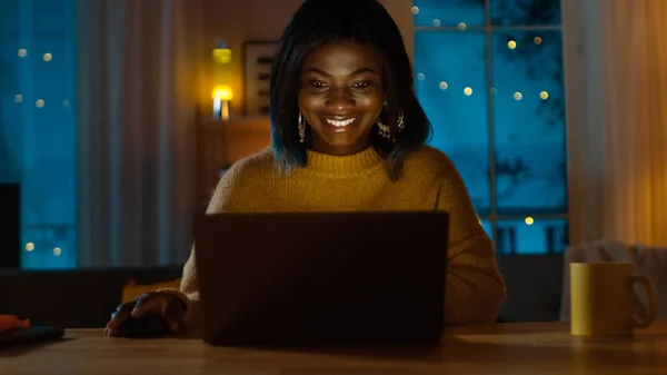 Retrato de una hermosa chica negra sonriente trabajando en una computadora portátil mientras está sentada en su escritorio en casa. Por la noche, la mujer creativa trabaja en una computadora en su acogedora sala de estar . —  Fotos de Stock