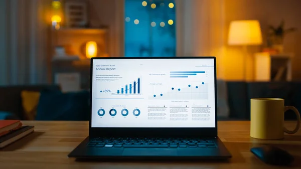 Laptop computer toont statistische infographics staat op een bureau in de woonkamer. Op de achtergrond gezellige woonkamer met warme verlichting op. — Stockfoto