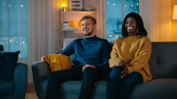 Boldog sokszínű fiatal pár néz komédia a TV-ben ülve a kanapén, hogy nevetni és élvezni show. Szép kaukázusi Boy és a Black Girl in Love költési idő együtt a Cozy apartmanban. — Stock Fotó