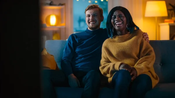 Boldog sokszínű fiatal pár néz komédia a TV-ben ülve a kanapén, hogy nevetni és élvezni show. Szép kaukázusi Boy és a Black Girl in Love költési idő együtt a Cozy apartmanban. — Stock Fotó