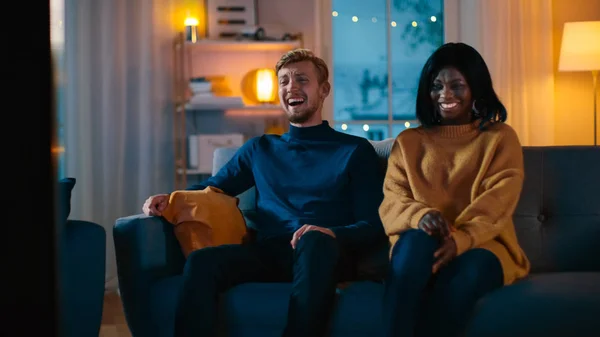 Boldog sokszínű fiatal pár néz komédia a TV-ben ülve a kanapén, hogy nevetni és élvezni show. Szép kaukázusi Boy és a Black Girl in Love költési idő együtt a Cozy apartmanban. — Stock Fotó