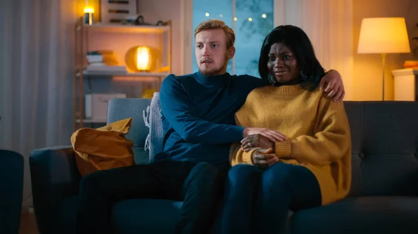 Happy Young Couple Watching Horror Movie on TV while Sitting on a Couch, they got Scared, Girl Covers eyes. Handsome Caucasian Boy and Black Girl in Love Spending Time Together. — Stock Photo, Image