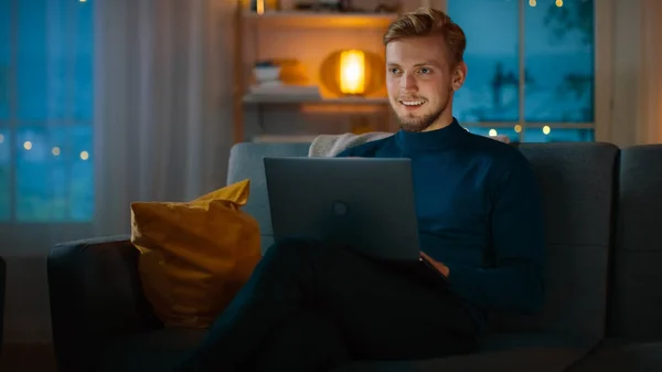 Το βράδυ όμορφος νέος άνθρωπος στο σπίτι κάθεται σε ένα καναπέ δουλεύει σε ένα φορητό υπολογιστή. Στο παρασκήνιο φιλόξενο σαλόνι. — Φωτογραφία Αρχείου
