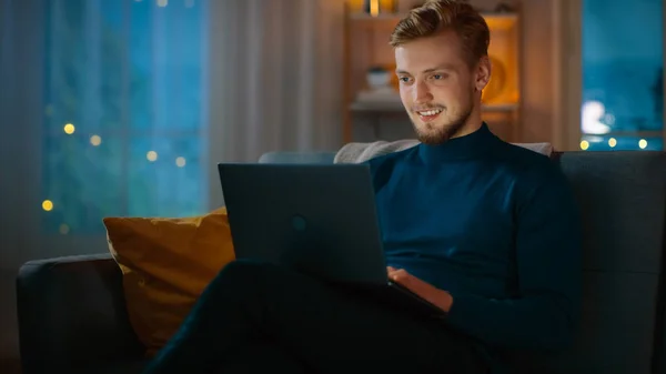 -Ban este csinos fiatal ember otthon ülő-ra egy dívány szerkezet-ra egy laptop számítógép. A háttérben hangulatos nappalijában. — Stock Fotó