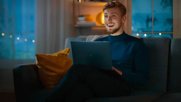Το βράδυ όμορφος νέος άνθρωπος στο σπίτι κάθεται σε ένα καναπέ δουλεύει σε ένα φορητό υπολογιστή και ρολόγια τηλεόραση κωμωδία και γέλια. — Φωτογραφία Αρχείου