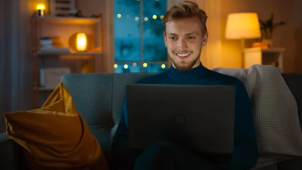 Το βράδυ όμορφος νέος άνθρωπος στο σπίτι κάθεται σε έναν καναπέ στο σαλόνι λειτουργεί σε ένα φορητό υπολογιστή. Χρησιμοποιεί τον υπολογιστή και περιηγείται μέσω του Διαδικτύου, έχει διασκέδαση και χαμόγελα. — Φωτογραφία Αρχείου