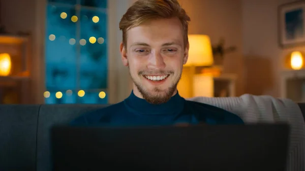 Na noite bonito jovem em casa sentado em um sofá na sala de estar funciona em um computador portátil. Ele usa o computador e navega através da Internet, se diverte e sorri . — Fotografia de Stock