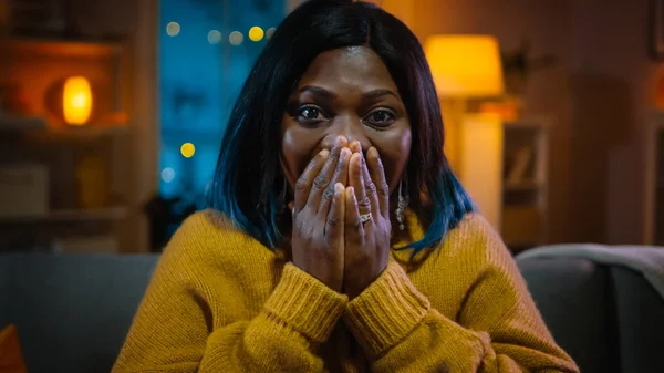 Retrato de una hermosa chica negra sentada en un sofá en casa por la noche, viendo películas en la televisión. Ella se pone realmente emocionada y emocional . — Foto de Stock