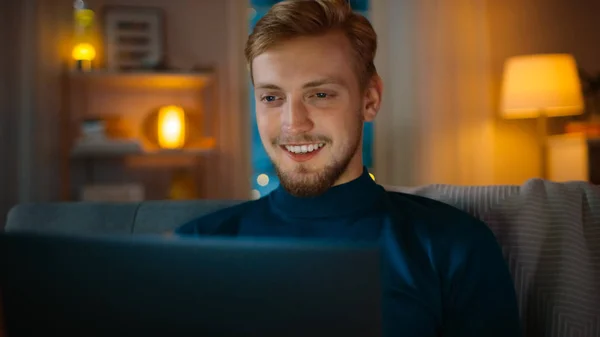 Na noite bonito jovem em casa sentado em um sofá na sala de estar funciona em um computador portátil. Ele usa o computador e navega através da Internet, se diverte e sorri . — Fotografia de Stock