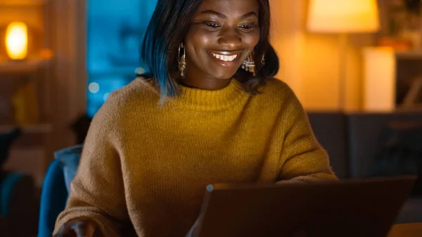 Retrato de una hermosa chica negra sonriente trabajando en una computadora portátil mientras está sentada en su escritorio en casa. Por la noche, la mujer creativa trabaja en una computadora en su acogedora sala de estar . —  Fotos de Stock