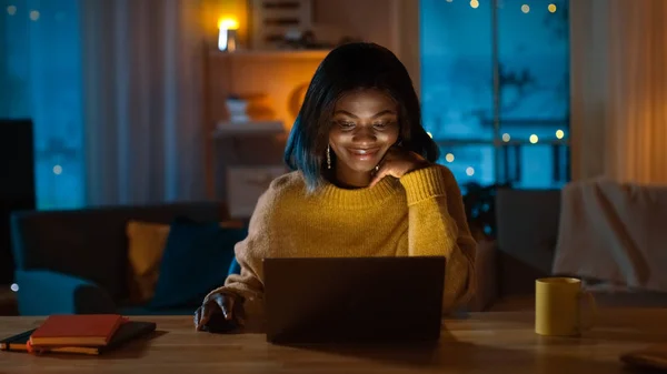 Retrato de la hermosa chica negra utiliza la computadora mientras está sentada en su escritorio en casa, lleva un suéter cálido. Por la noche, la mujer creativa trabaja en una computadora en su acogedora sala de estar . —  Fotos de Stock