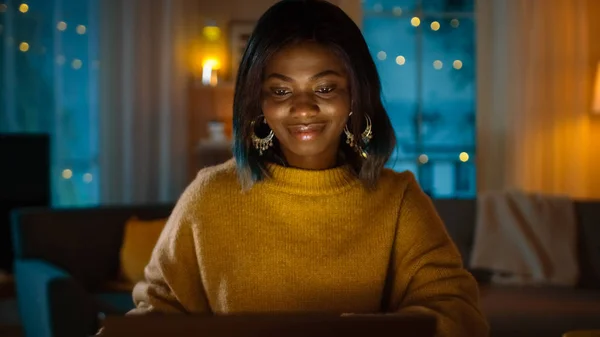 Portrét krásné černé dívky pracovat na notebooku při sezení u jejího stolu doma. V večerní tvůrčí ženě pracují v počítači v útulném obývacím pokoji. — Stock fotografie