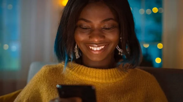 Portrét krásné černé dívky používá smartphone při sezení na pohovce doma, její tvář je osvětlená obrazovkou. V rozkošné ženě úsměvů a procházky přes Internet. — Stock fotografie