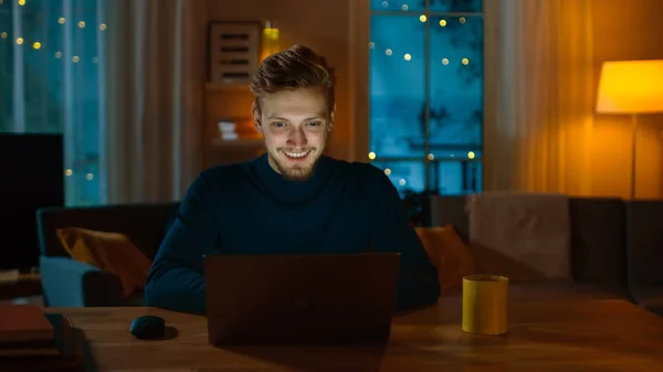 Όμορφος χαμογελαστός άνθρωπος δουλεύει σε ένα φορητό υπολογιστή ενώ κάθεται στο γραφείο του στο σπίτι. Νέος ελεύθερος επαγγελματίας εργάζεται στον υπολογιστή στο φιλόξενο σαλόνι του με ζεστό βραδινό φωτισμό ενεργοποιημένος. — Φωτογραφία Αρχείου