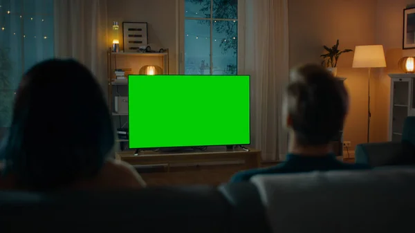 Oturma odasında bir kanepede otururken birkaç saatler yeşil mock-up ekran TV. Erkek arkadaş ve kız arkadaşı için romantik akşam. — Stok fotoğraf